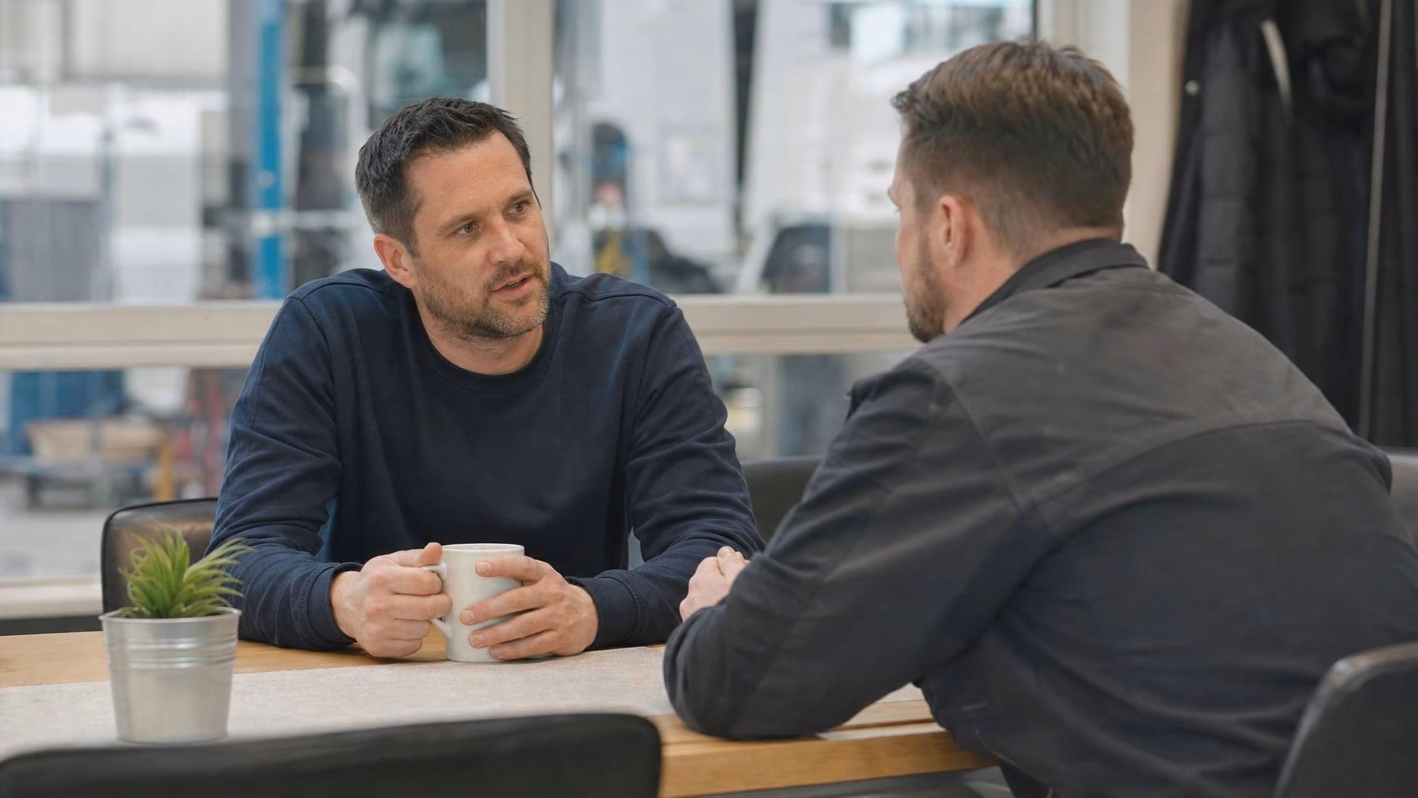 Disponent im Halbprofil im Gespräch mit Fahrer in Werkstattjacke am Pausenraum-Tisch, Kaffeetasse