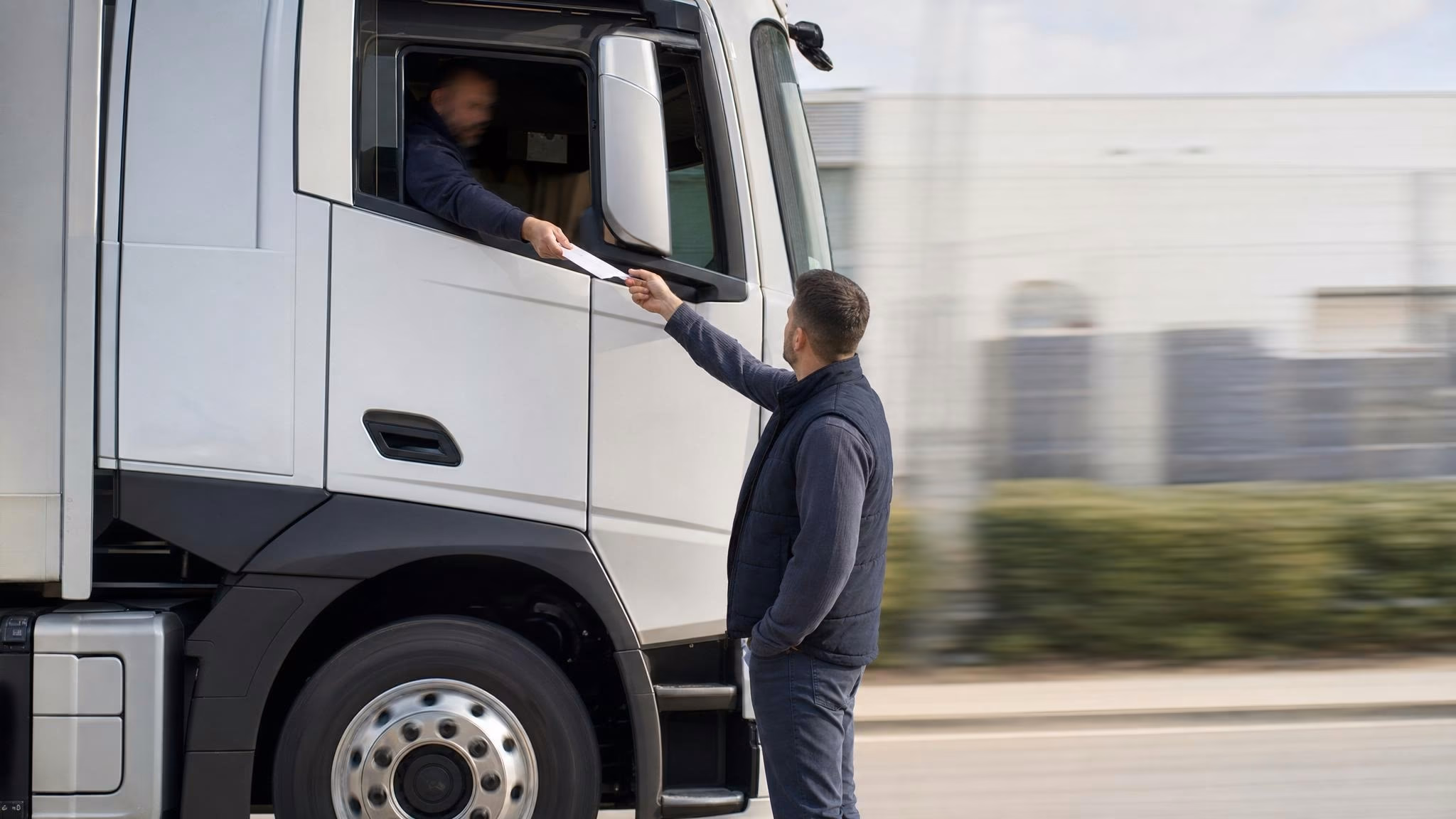 Disponent mit Klemmbrett am offenen Lkw-Cockpit, Fahrer reicht im Halbprofil ein Blatt Papier ohne lesbaren Text herüber, dunkelblaue Coveralls