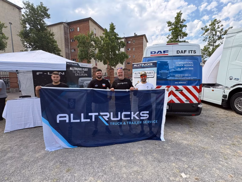 Czterech członków zespołów Alltrucks i Nordest Trucks trzyma duży baner Alltrucks przed pojazdem serwisowym DAF-ITS na stoisku podczas 12. edycji Concentración de Camiones w Olot