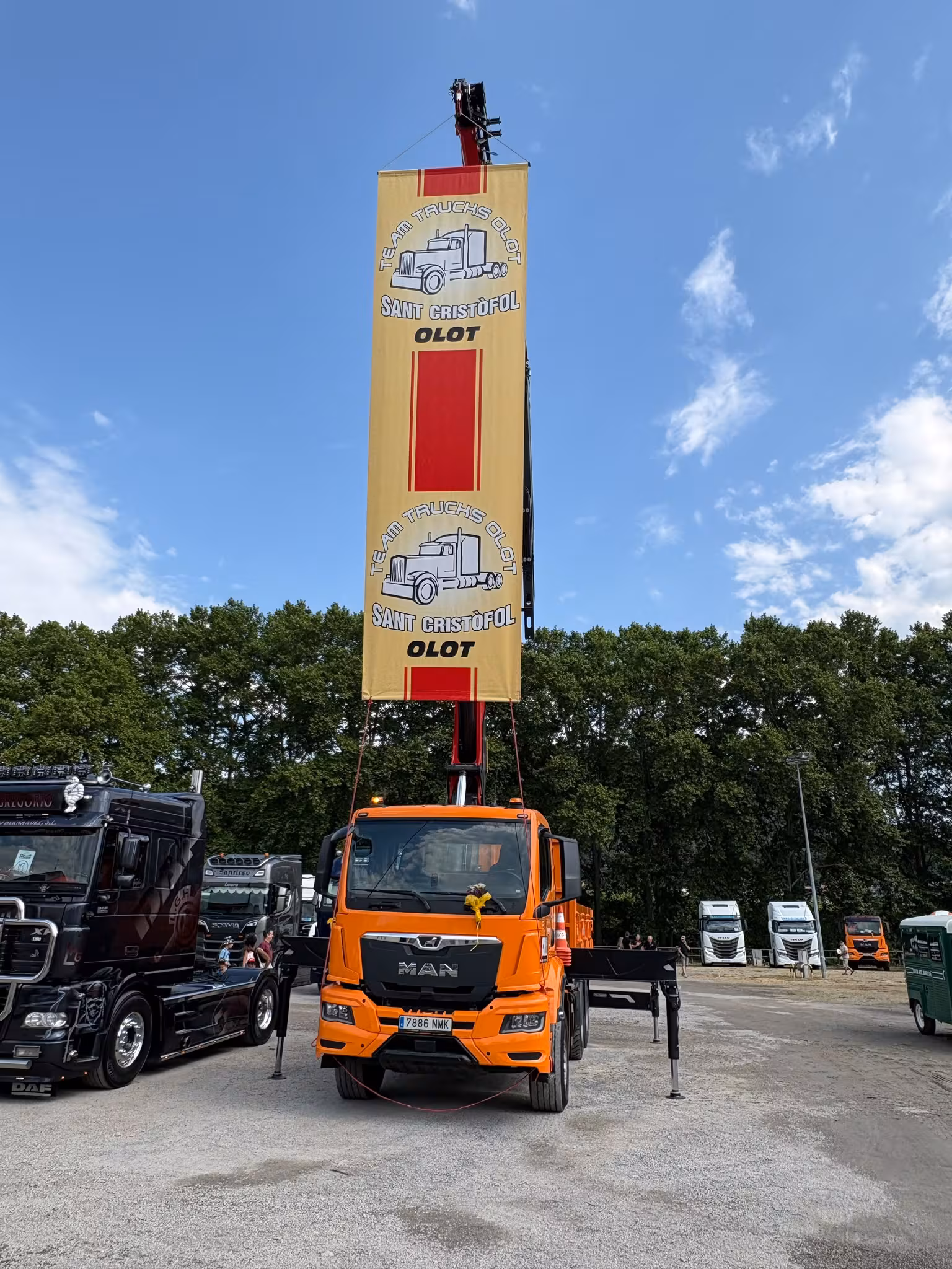 Oranžinės spalvos MAN sunkvežimio krovimo kranas aukštai virš renginio teritorijos Olote pakelia „Team Truck Olot“ vėliavą su užrašu „Sant Cristòfol“