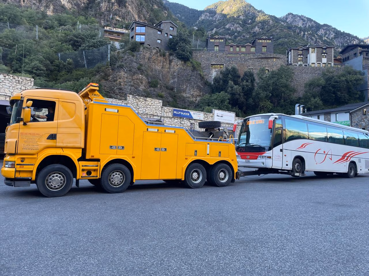 En gul tungtransportbilsbärgare från SERGRU bogserar en vit turistbuss över en parkeringsplats i Andorra, med bergen i bakgrunden