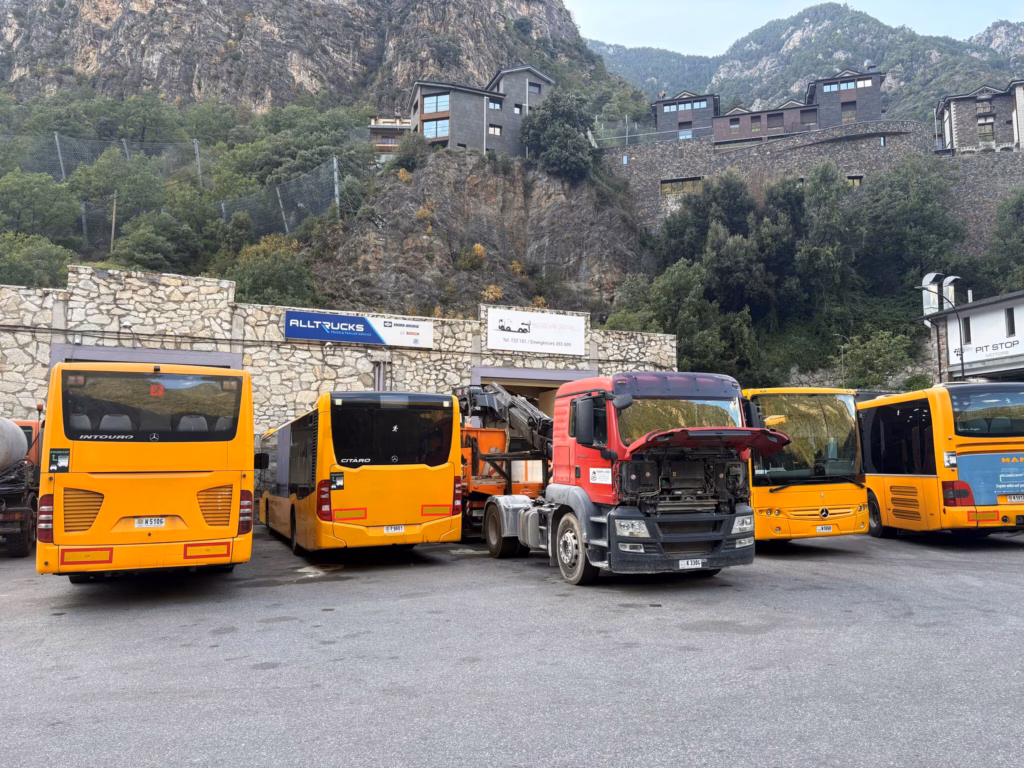 Delante del taller de SERGRU en Andorra hay autobuses de línea amarillos y un camión tractor rojo; detrás, una pared rocosa y las montañas
