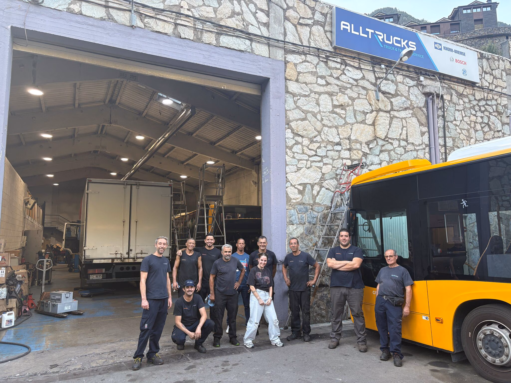 Elva medarbetare från SERGRU står framför verkstadens port; på naturstensväggen hänger Alltrucks-skylten, och till höger står en gul buss