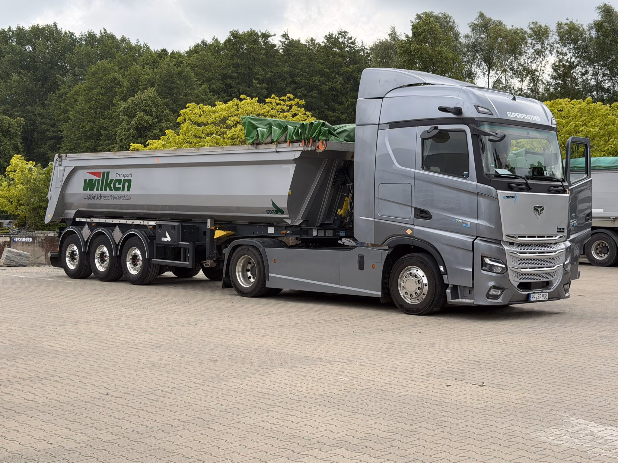 SuperPanther eTopas 600 con un semirremolque basculante cargado de la empresa de transportes Wilken, cubierto con una lona verde, en un patio empedrado