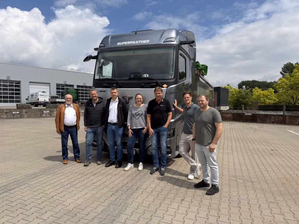 Siete participantes en la reunión de socios de Alltrucks posan delante del tractor de remolque plateado SuperPanther eTopas 600 en el patio de Melle