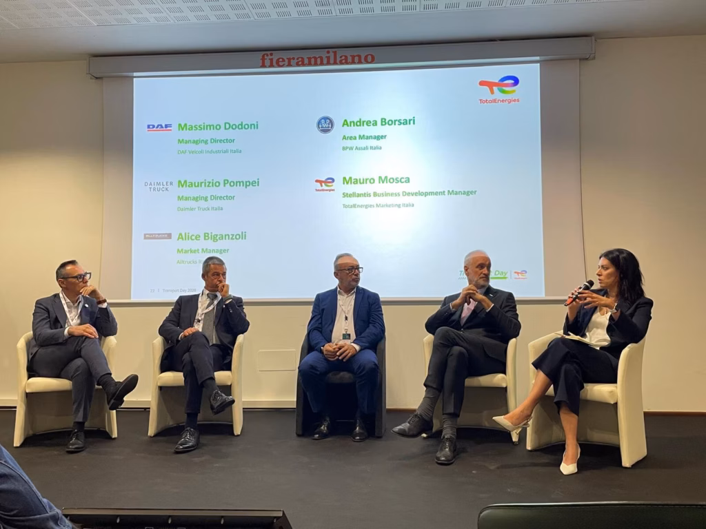 Cinco invitados en el podio de la mesa redonda del Transport Day 2026 de TotalEnergies en Milán; detrás, la pantalla con los nombres de los participantes de DAF, Daimler Truck, BPW, TotalEnergies y Alltrucks