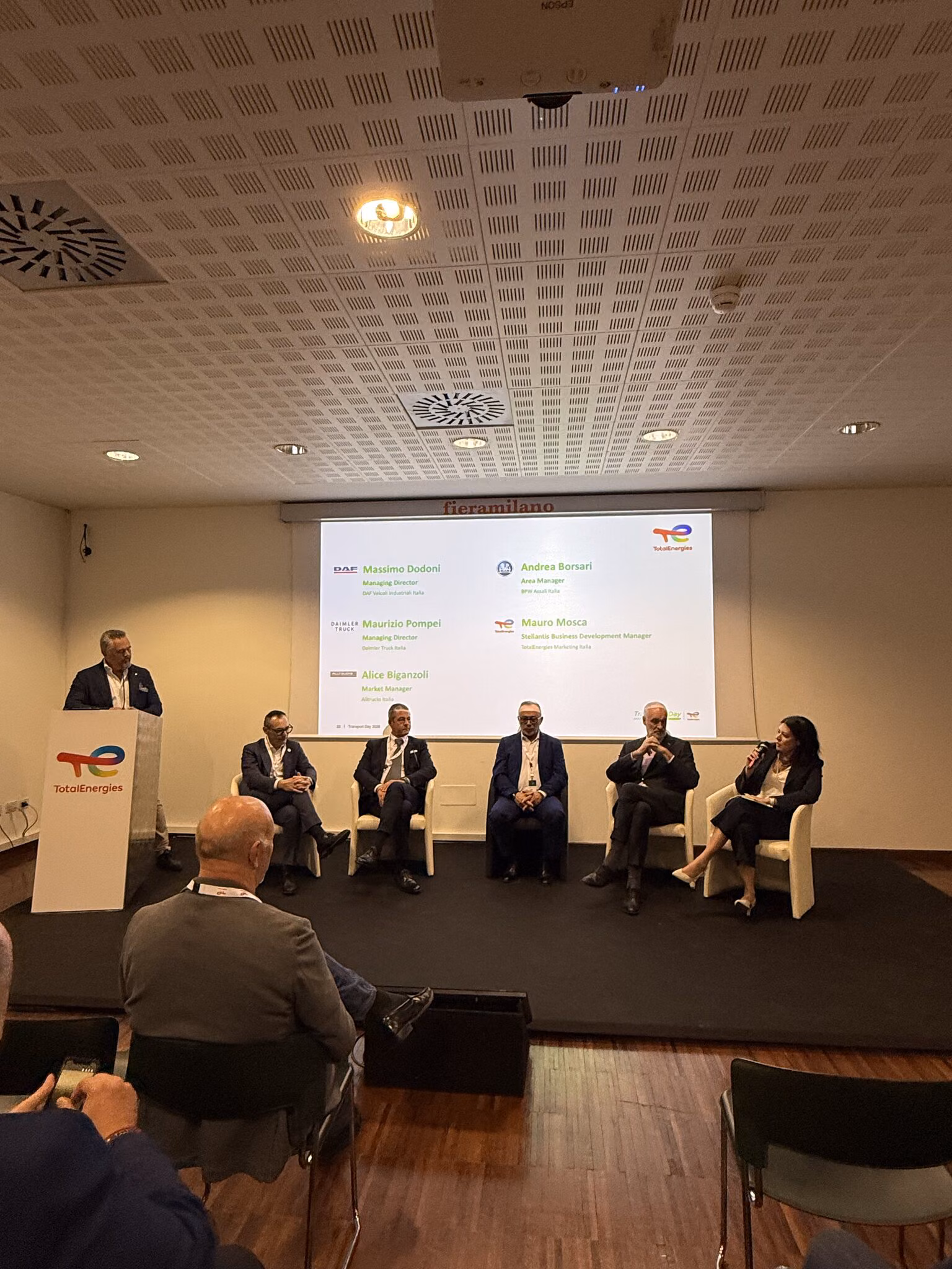 Sala la Transport Day 2026 din Milano, cu public, un moderator la pupitrul TotalEnergies și cinci invitați la masa rotundă în fața ecranului